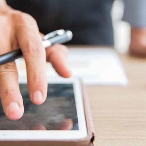 Close-up of hand of unrecognizable businessman standing at table, holding pen and using digital tablet in office